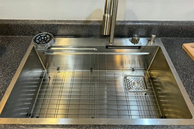 Stainless steel kitchen sink with a rack inside, faucet, and soap dispenser, on a dark countertop.
