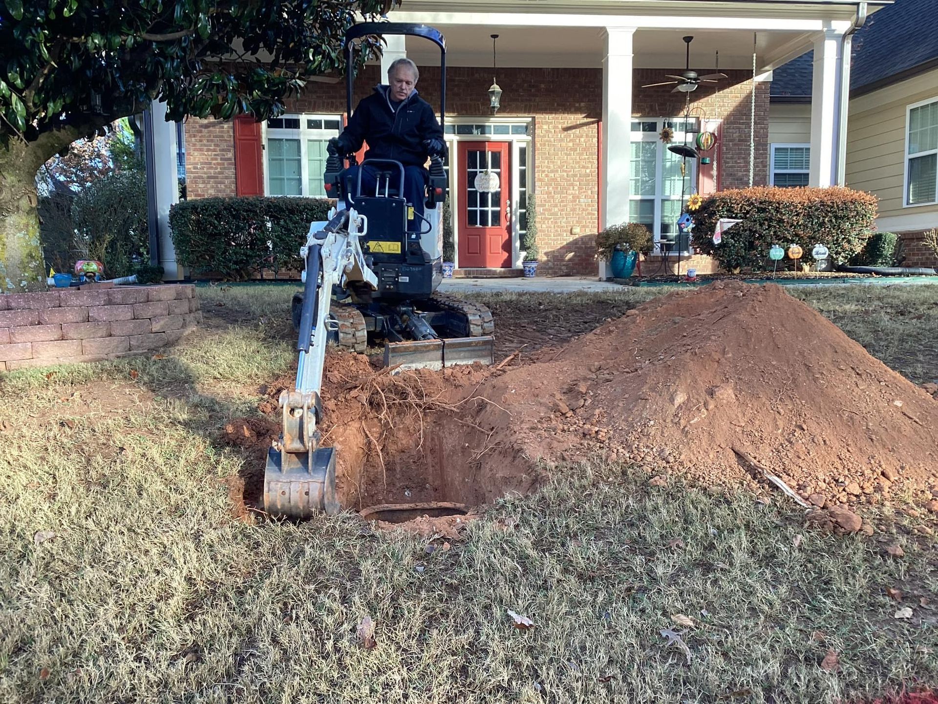 A man is using an excavator to dig a hole in the ground in front of a house.