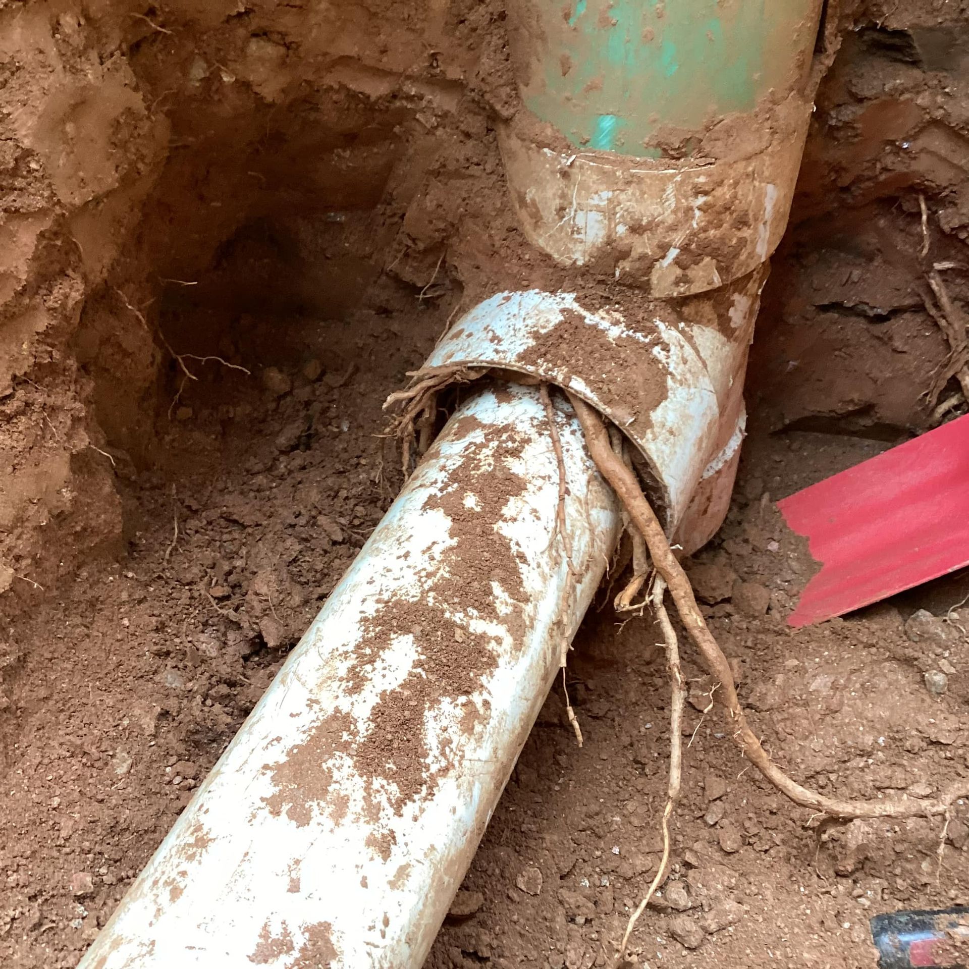 A white pipe is laying in the dirt next to a red shovel.