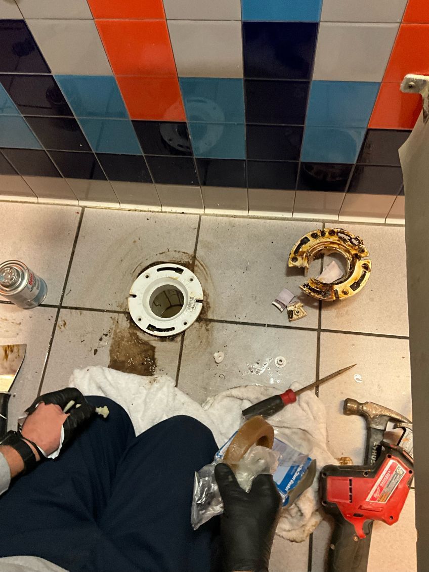 Person repairing a toilet: tools and parts on the floor, colorful tiled walls, dark stains around the drain.