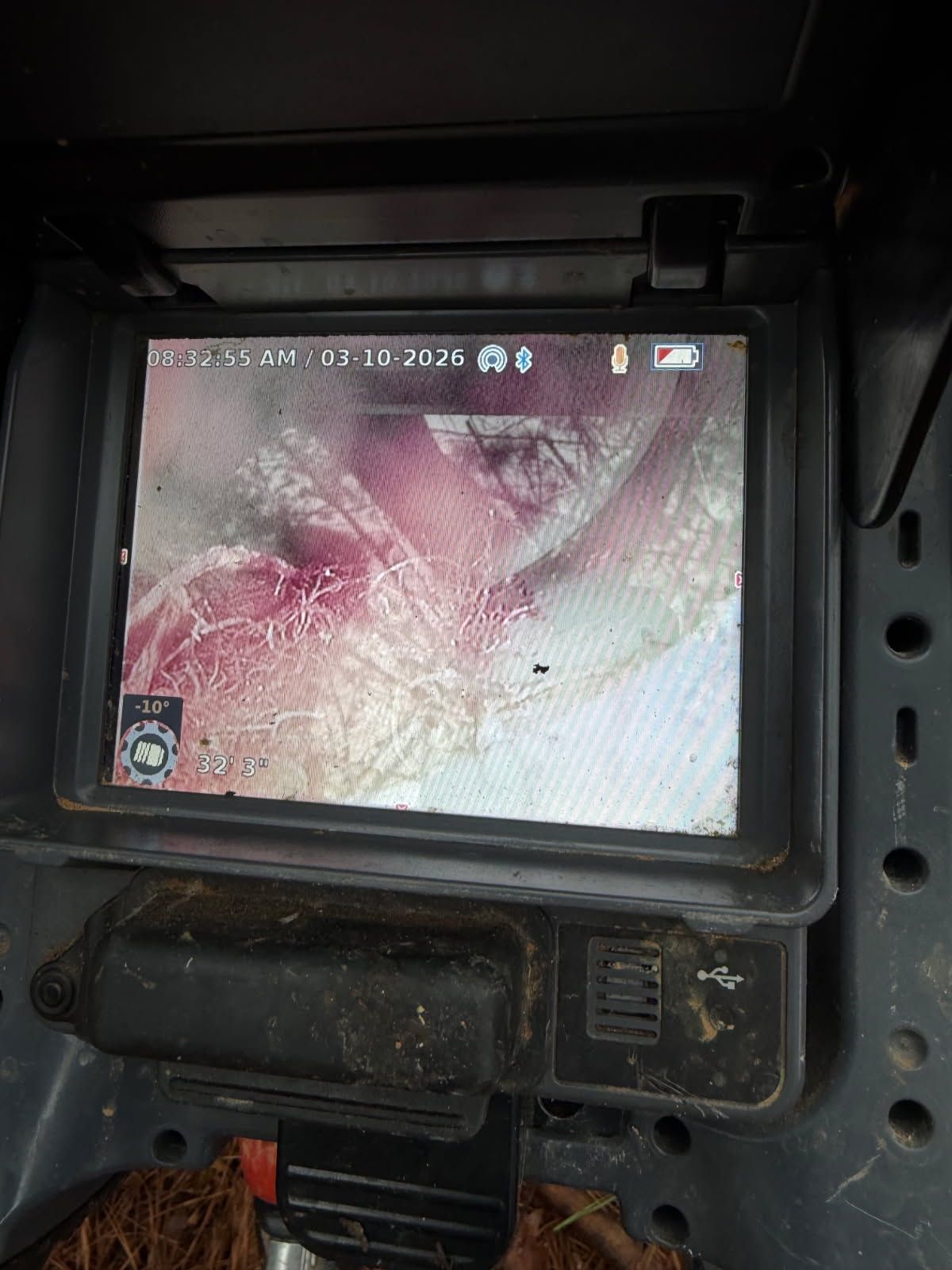 A handheld sewer camera monitor displays an underground pipe inspection with visible roots and debris.