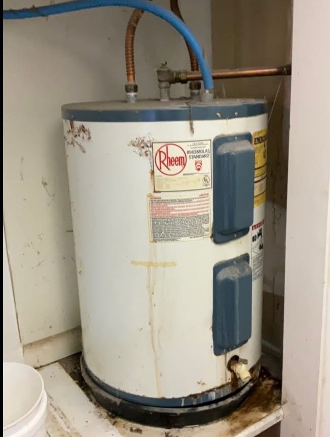 Rheem electric water heater in a utility closet with corrosion and water damage at the base.
