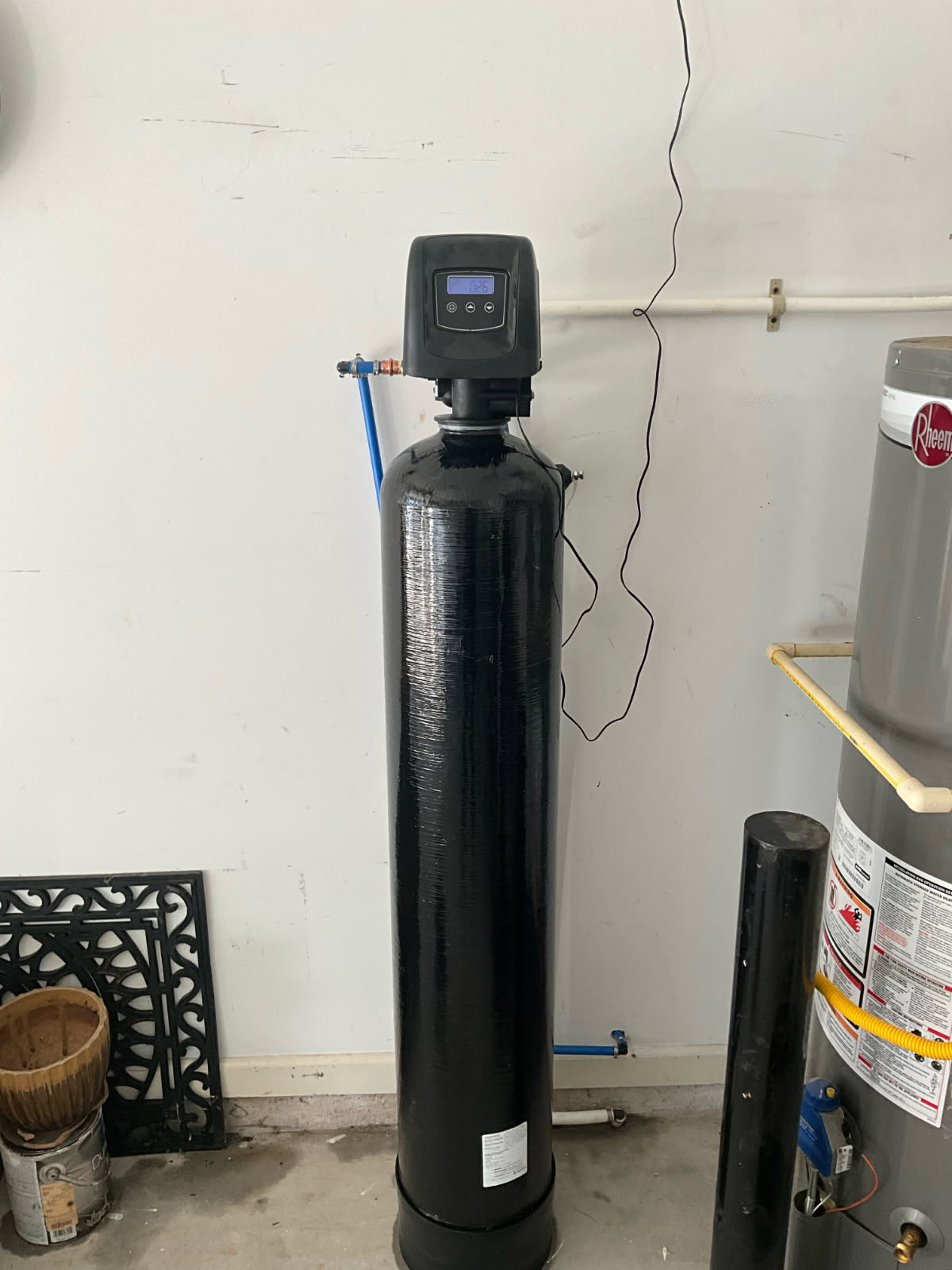 Black water softener tank with digital display, installed in a garage next to a water heater.