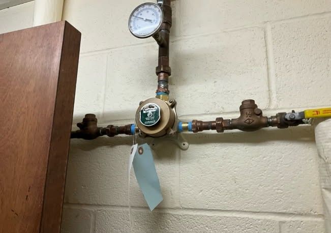 Plumbing with a pressure gauge and valve system on a brick wall. A blue tag hangs from the assembly.