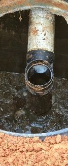 A pipe extends into an open, dirt-filled septic or drainage access pit containing dark liquid and debris.