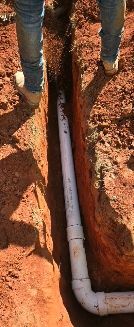 A person stands on either side of a narrow dirt trench containing a white PVC pipe with a 90-degree elbow joint.