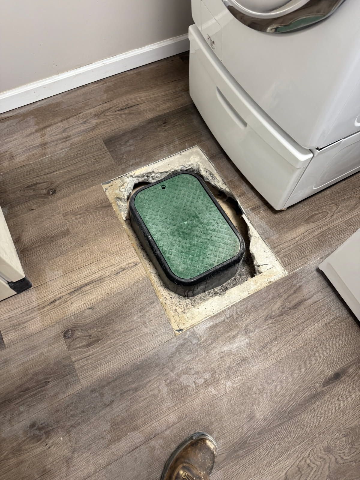 Green irrigation valve box in a floor opening, near a white appliance on wood-look flooring.