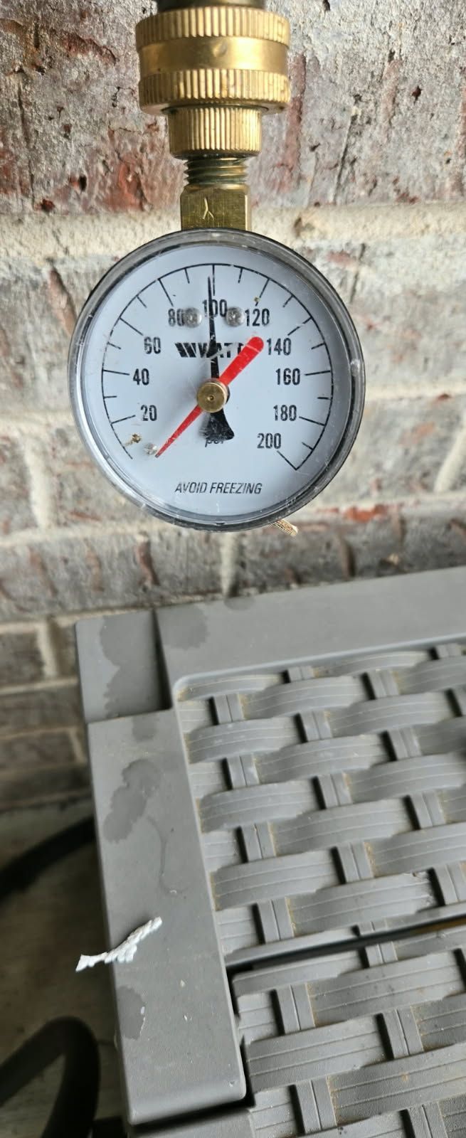 A pressure gauge showing about 100 PSI, attached to a brass fitting on a brick wall.
