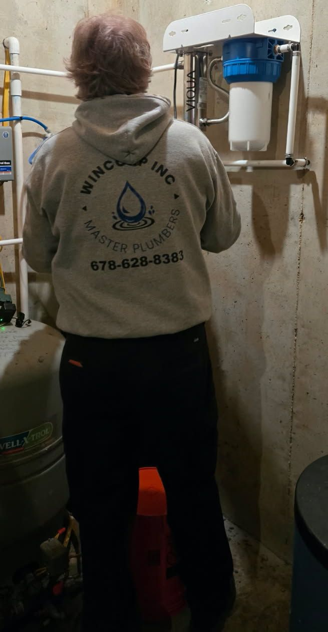 A technician in a gray hoodie works on a wall-mounted blue water filtration system in a concrete basement.