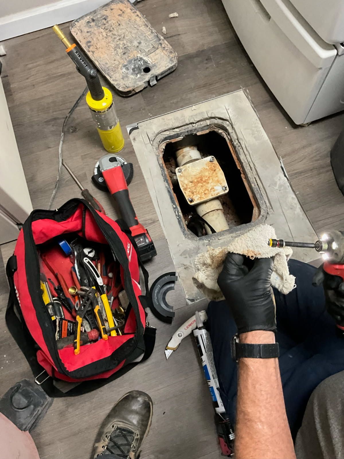 Person in black gloves repairs plumbing with tools, near a toolbox and appliance.
