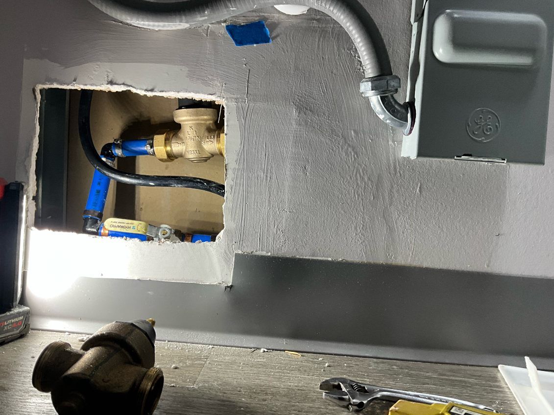 Plumbing and electrical components visible within an access opening, including pipes, valves, and a metal box.