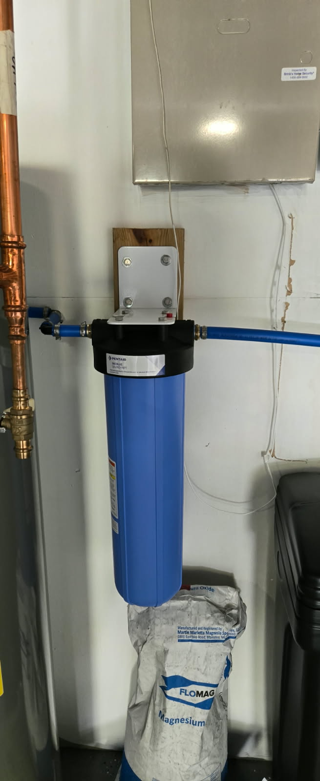A blue water filter installed on a white wall. Copper pipes and a bag of filter media are also visible.