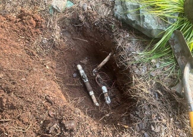 Hole dug in dirt reveals buried pipes, near shovel and grass.