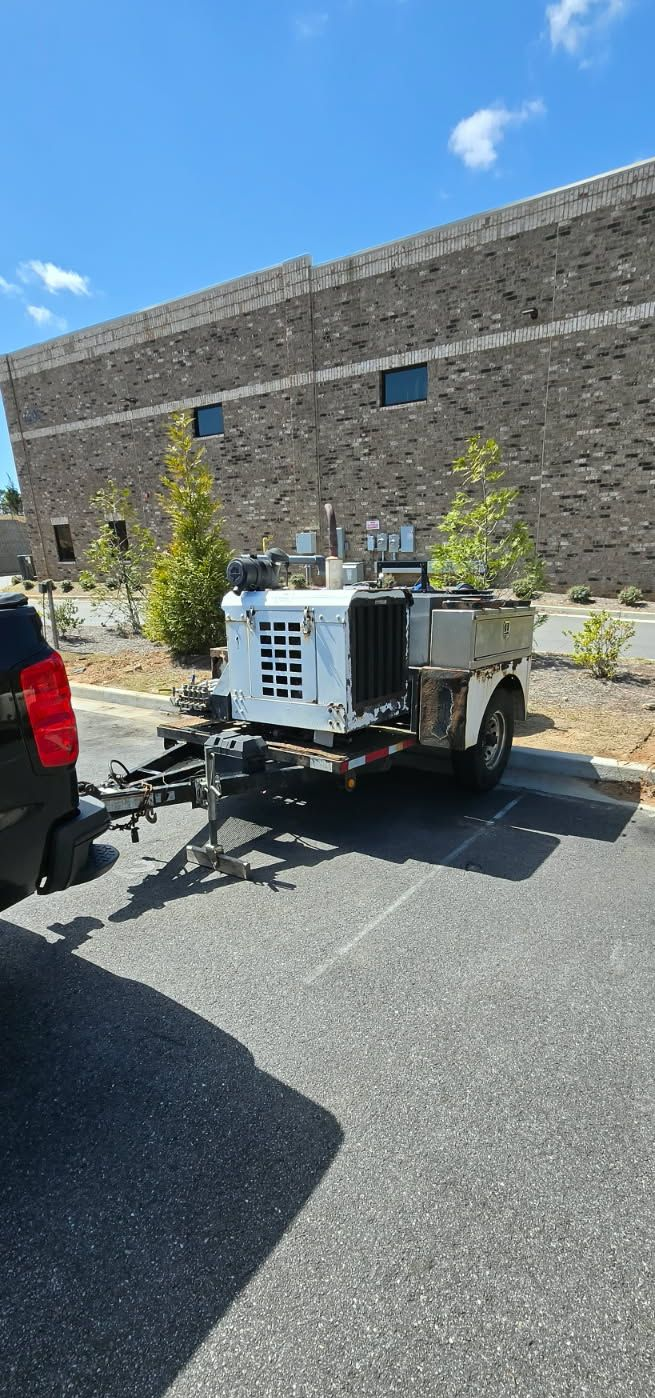 A white industrial generator on a trailer hitched to a vehicle in a parking lot in front of a stone building.