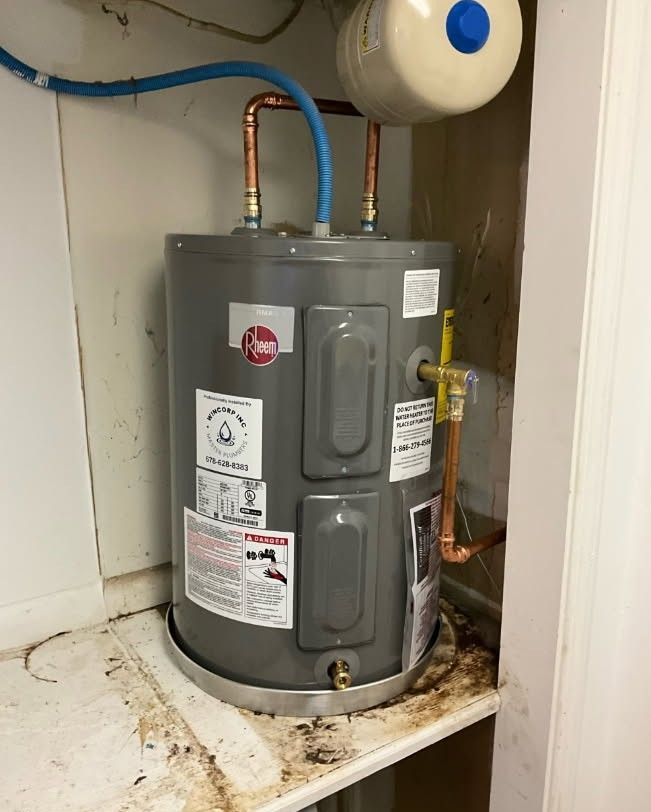 Gray water heater in a closet, with copper pipes and a white expansion tank. Brown water stains around the base.