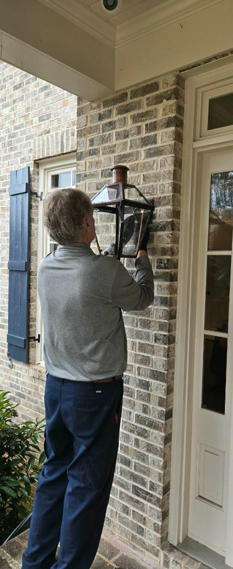 A person standing on a porch is working on a wall-mounted light fixture.
