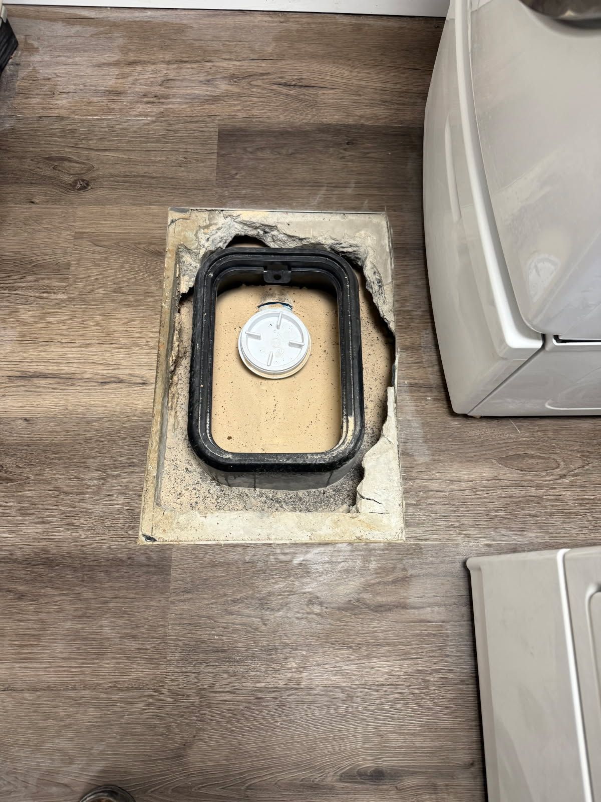 Rectangular laundry room floor access panel with a black seal and a white gauge.