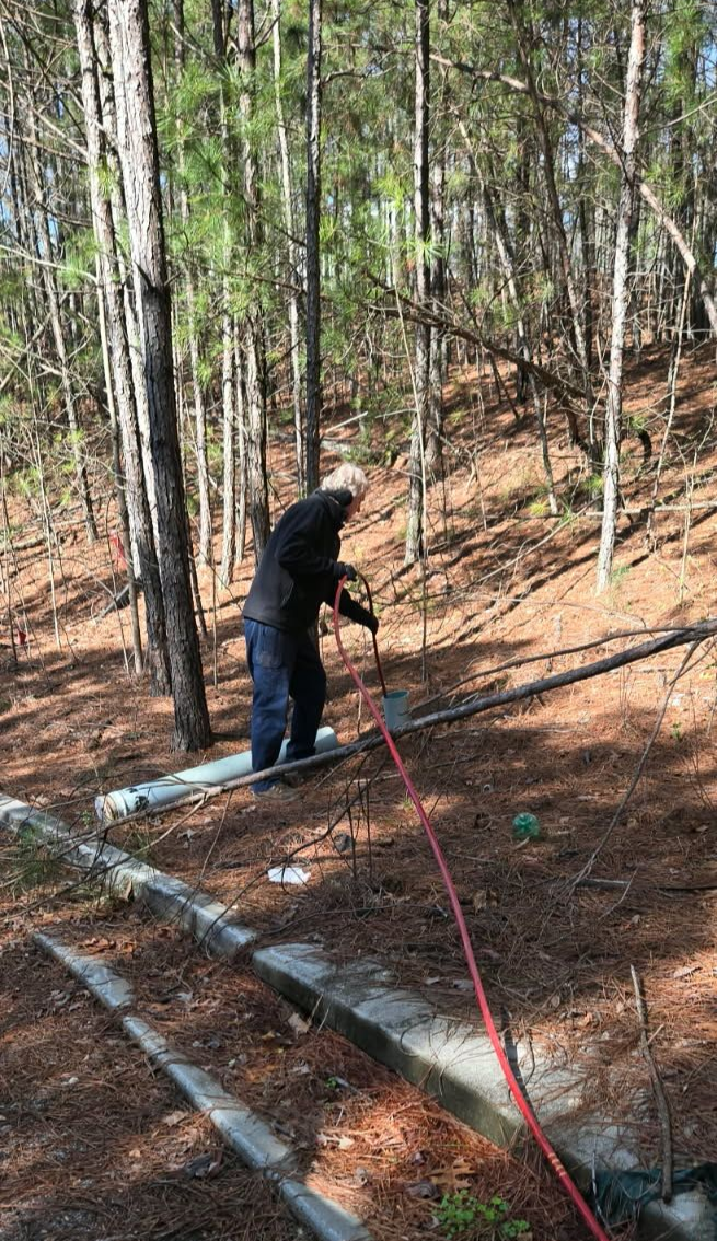 A person works with a long, narrow metal frame or pipes set in a wooded area with pine needles on the ground.