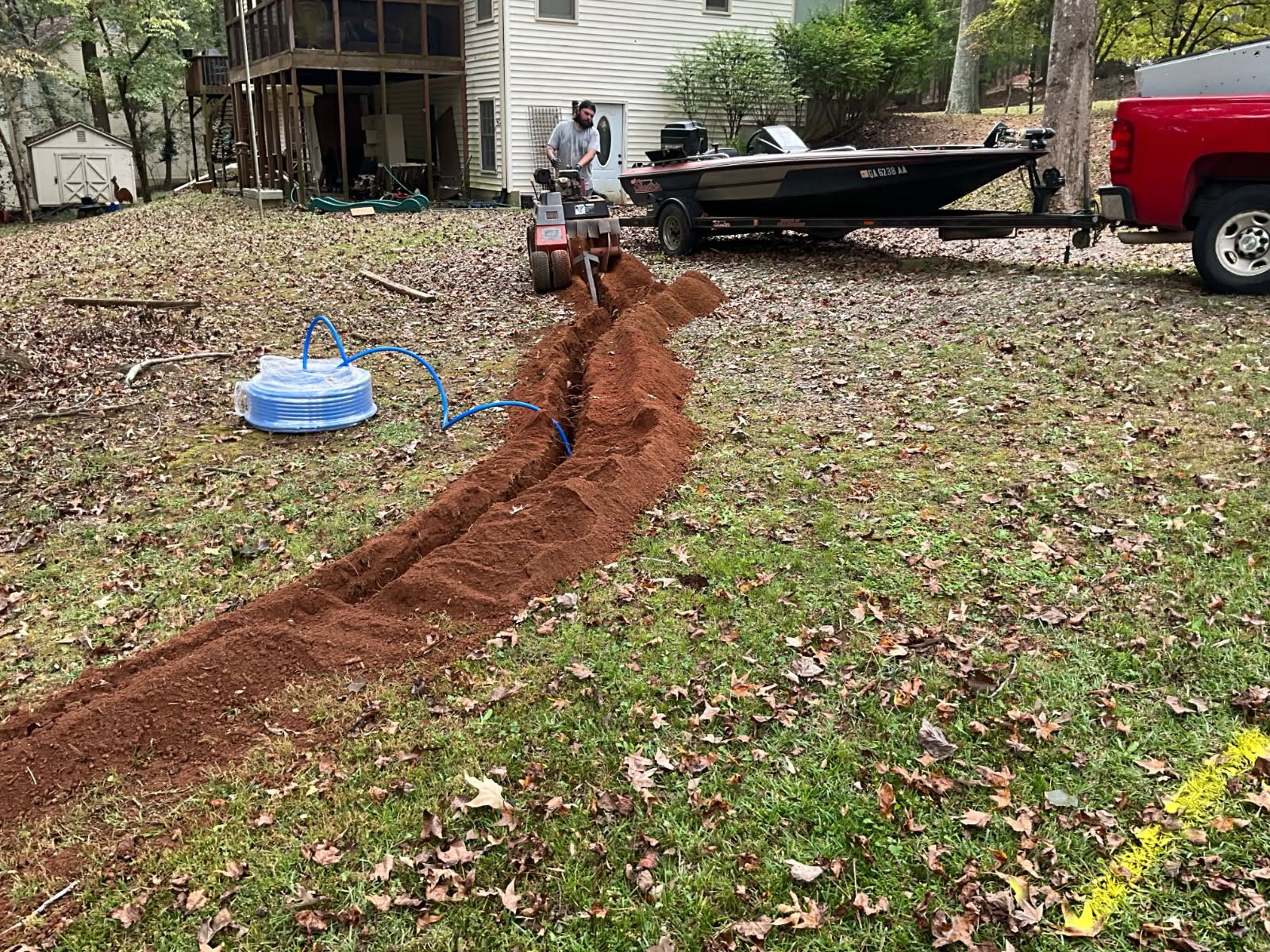 Man using a trencher, creating a narrow ditch in a grassy yard. Boat and truck are nearby.
