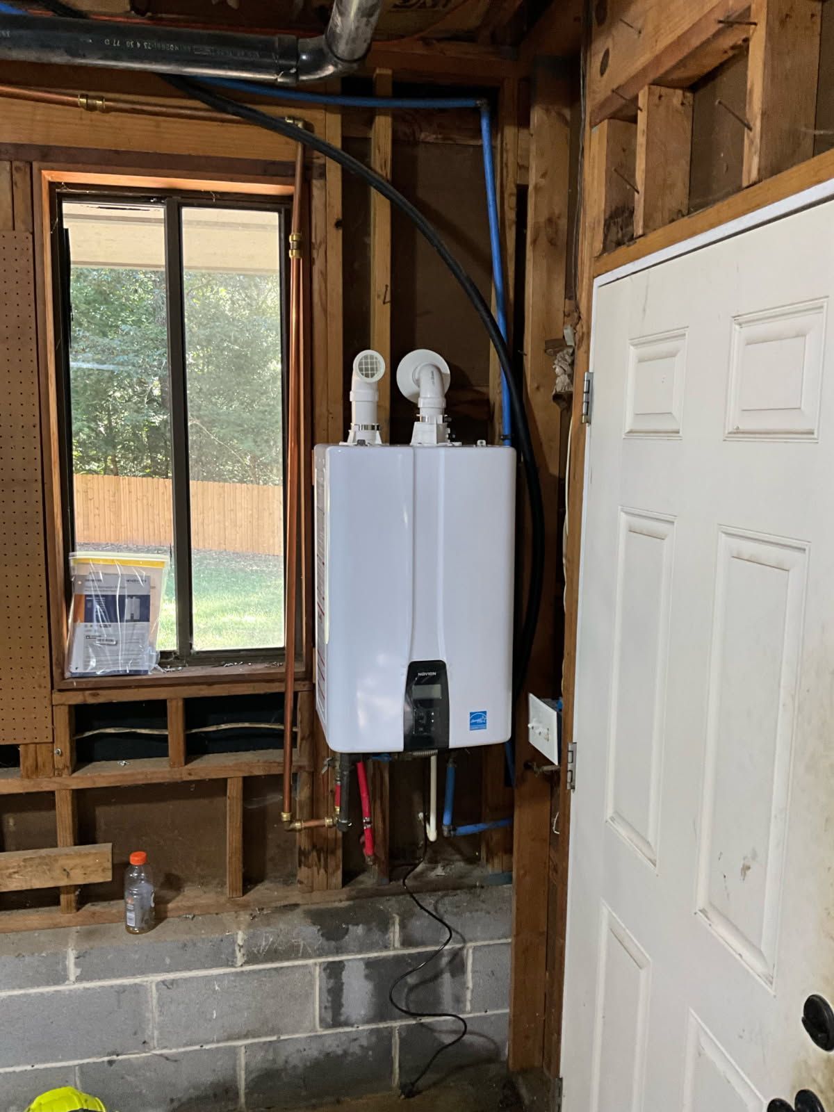 Gas water heater installation in a room with exposed studs and a window.