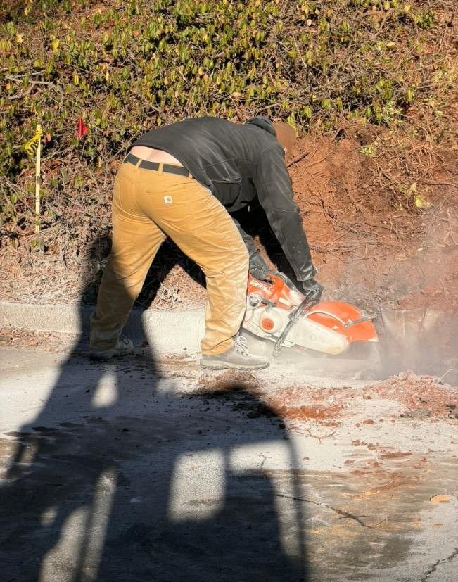Person in tan pants and black jacket cutting concrete with a power saw outdoors, with the sun casting a shadow.