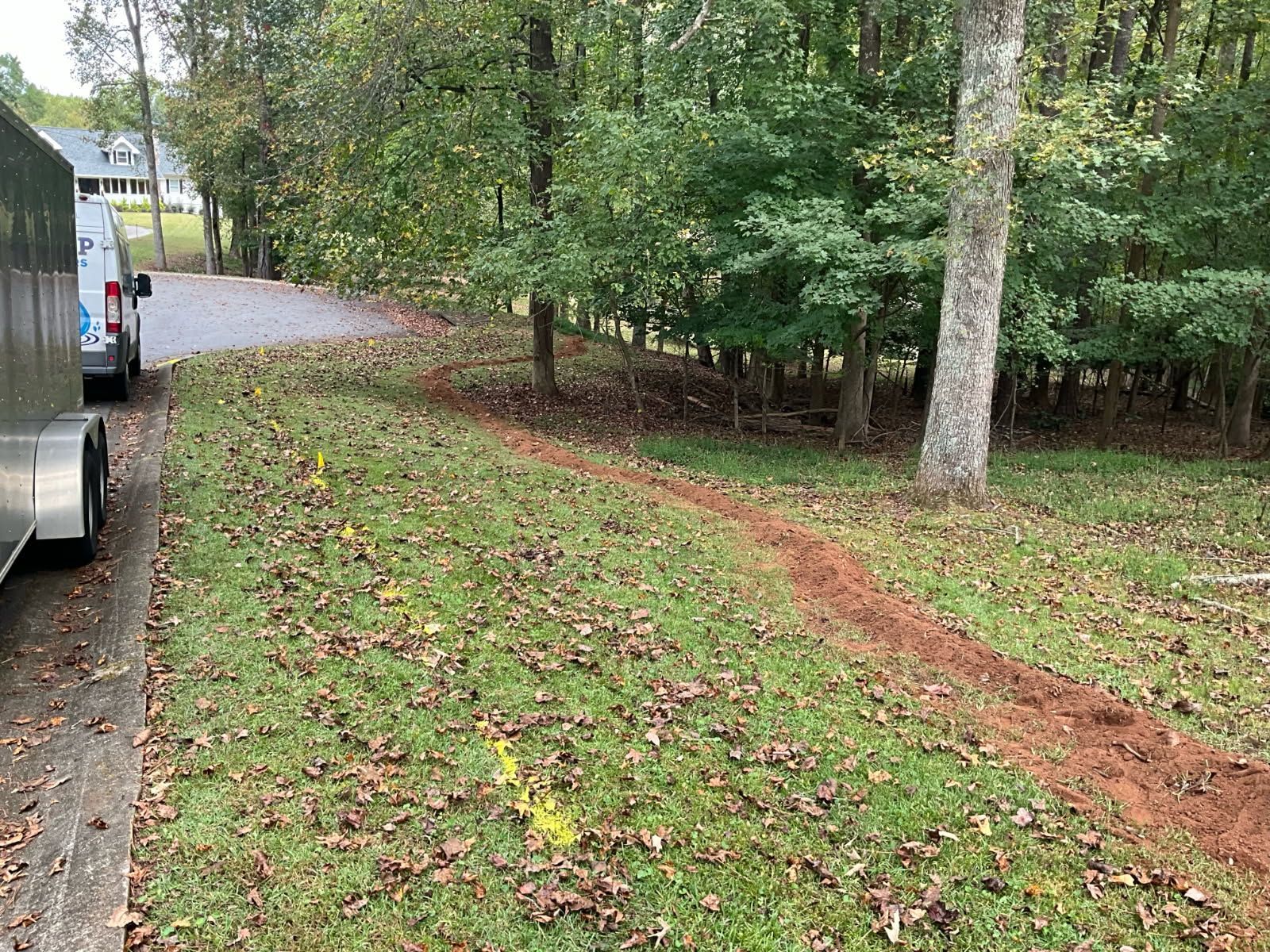 A trench dug in a grassy yard, near a road and trees, with a utility trailer parked on the side.