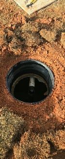 An open, circular irrigation valve box set into dirt, with a single white PVC pipe fitting visible at the bottom.