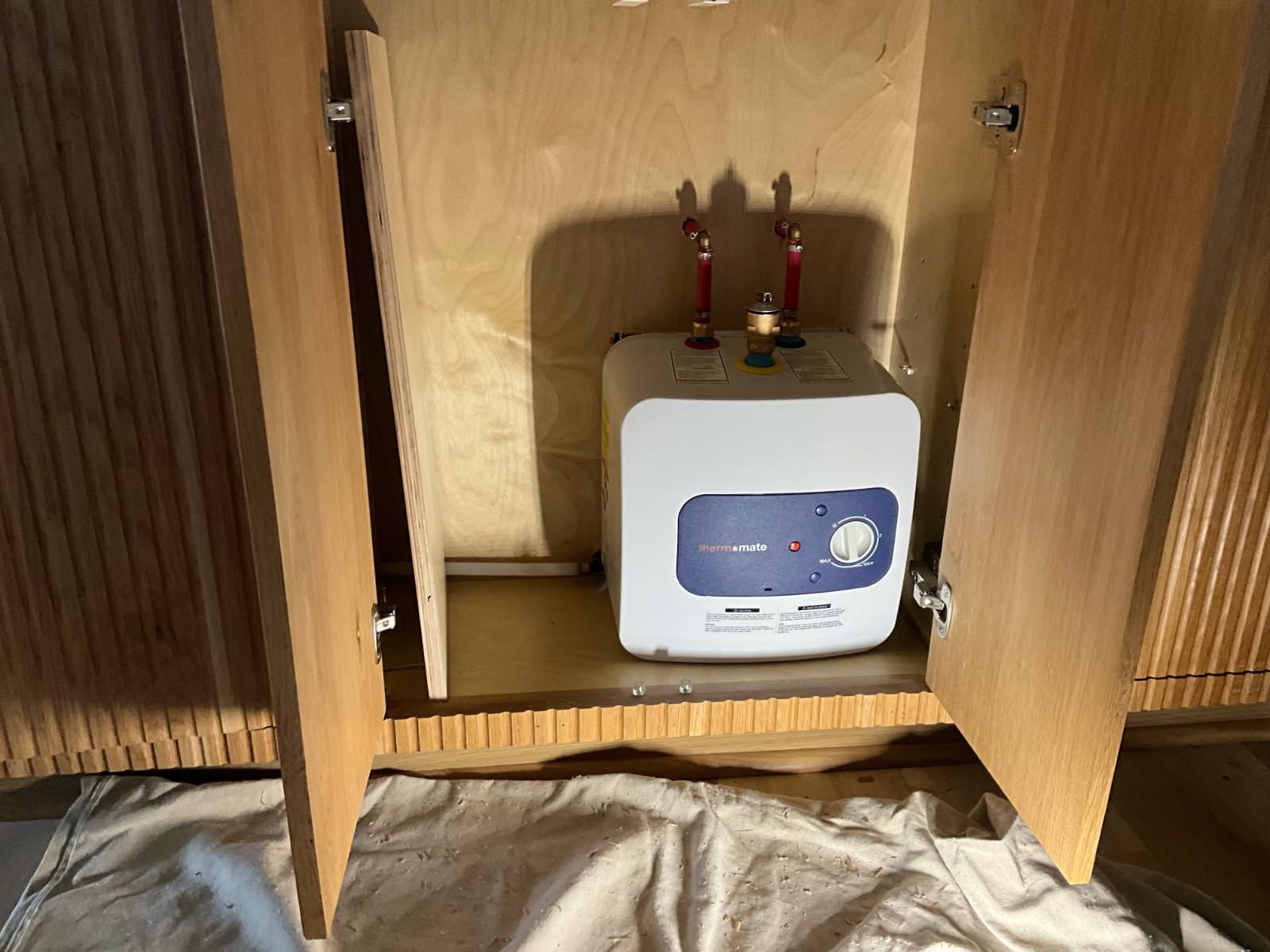 Small water heater inside a wooden cabinet with open doors; pipes visible.