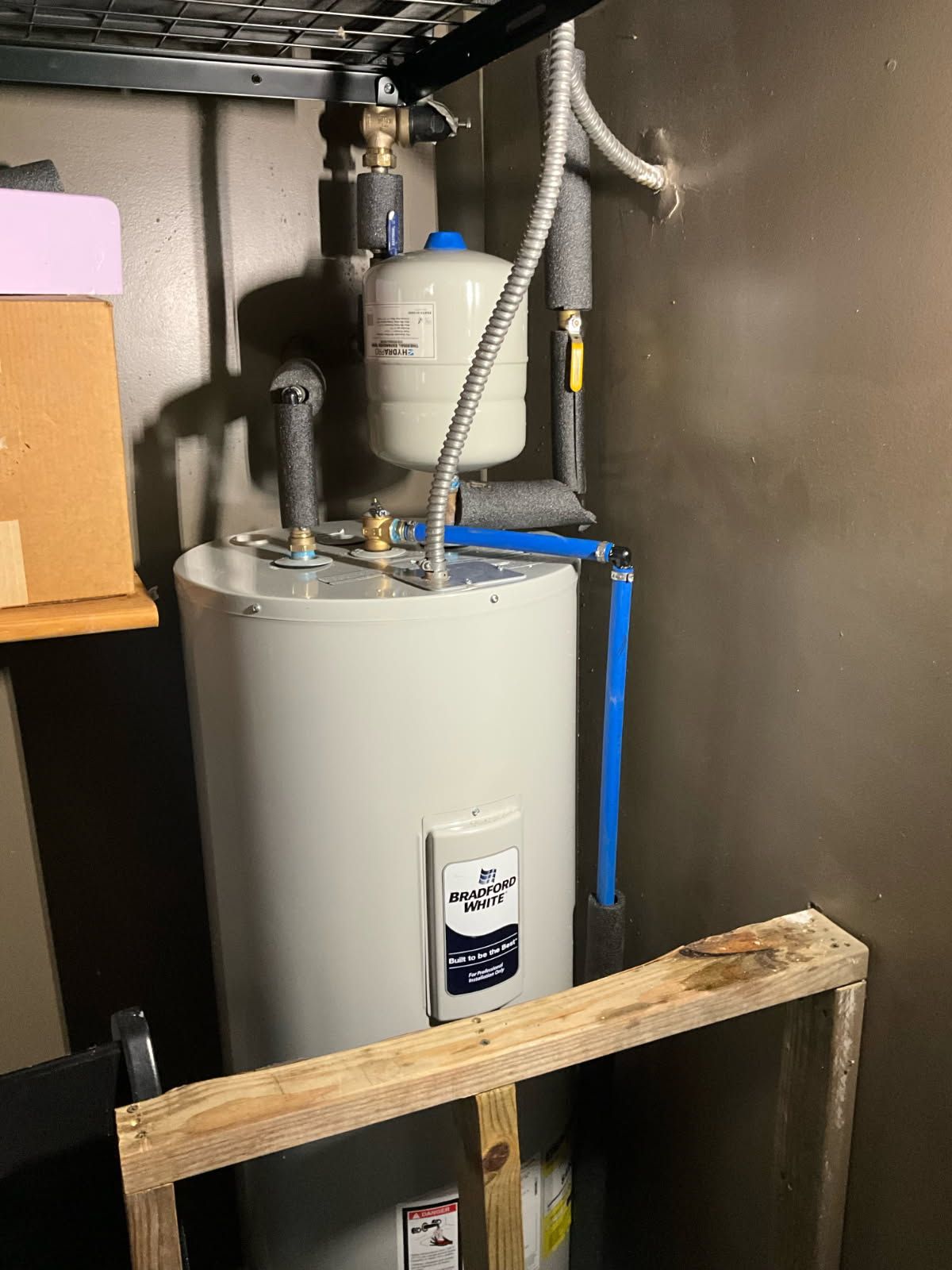 A tall, cylindrical water heater in a basement.  Blue and silver pipes connect to the tank.