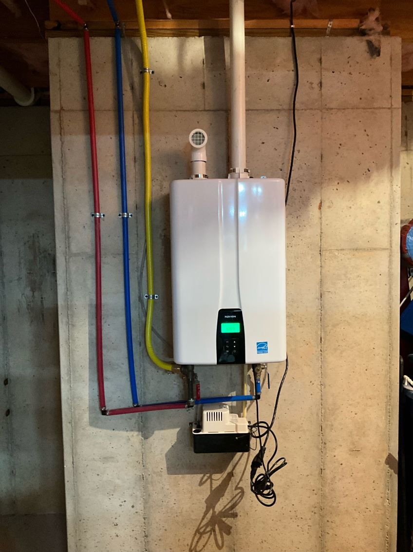 White tankless water heater mounted on a concrete wall with red, blue, and yellow pipes.