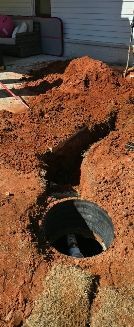 A vertical excavation trench with a dark circular access point revealing a white pipe in the red clay dirt near a house.
