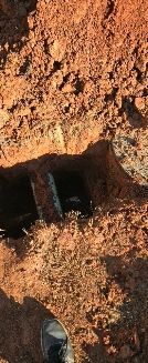 A view from above showing a deep, dark hole in the reddish-brown earth with a person's shoe visible at the bottom.