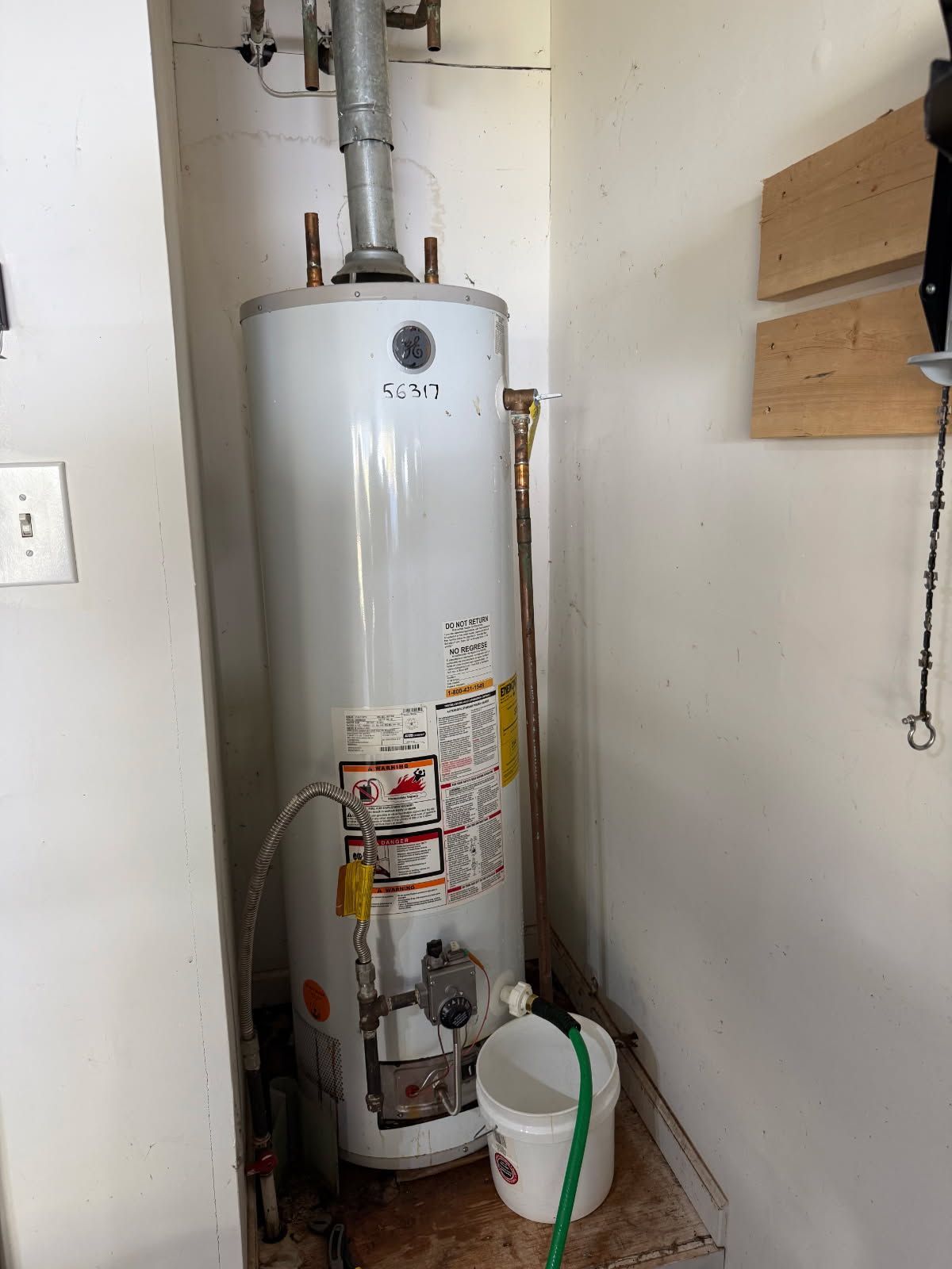 Water heater in a utility closet with vent pipe and attached water lines. White bucket is on the floor.