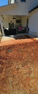 A patio with outdoor furniture beneath a covered porch, fronted by a yard of red soil and dry, patchy grass.