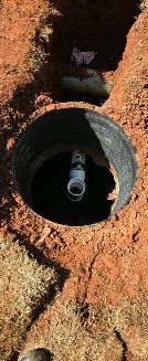 A vertical view of an uncovered irrigation valve box in a trench of red dirt with a visible white PVC pipe and fitting.