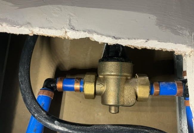 Brass pressure reducing valve with blue PEX tubing, copper crimp rings, and black insulated wire in a wall.