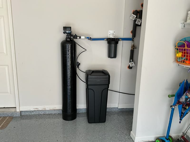 Water filtration system in a garage setting; black tanks and pipes against a white wall.