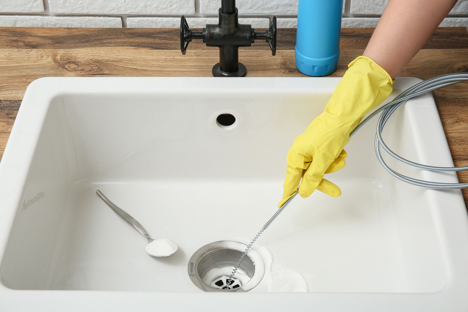Person wearing yellow gloves cleans a white sink drain with a snake tool, cleaning supplies nearby.