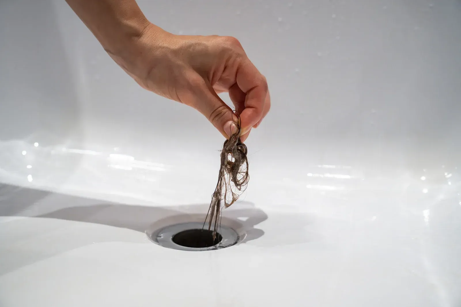 Hand holding a clump of hair above a bathroom drain. The drain is in a white tub.