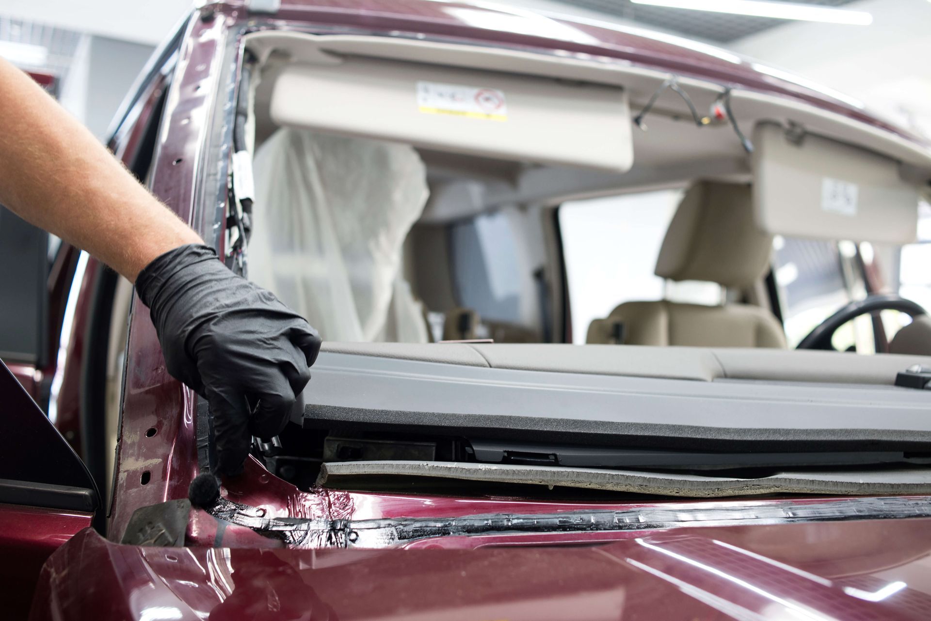 Gloved hand removing windshield from a red car in a repair shop; interior visible.