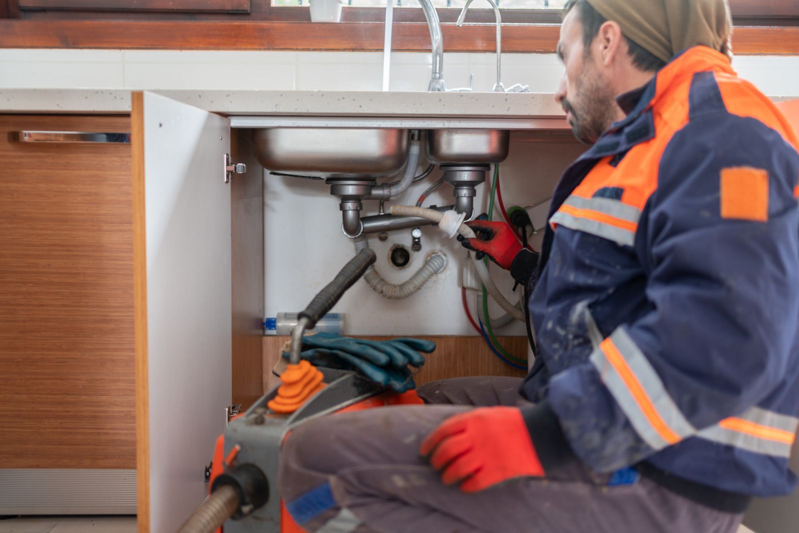 Plumber repairs kitchen sink pipes. Blue and orange work jacket, red gloves, inside cabinet.
