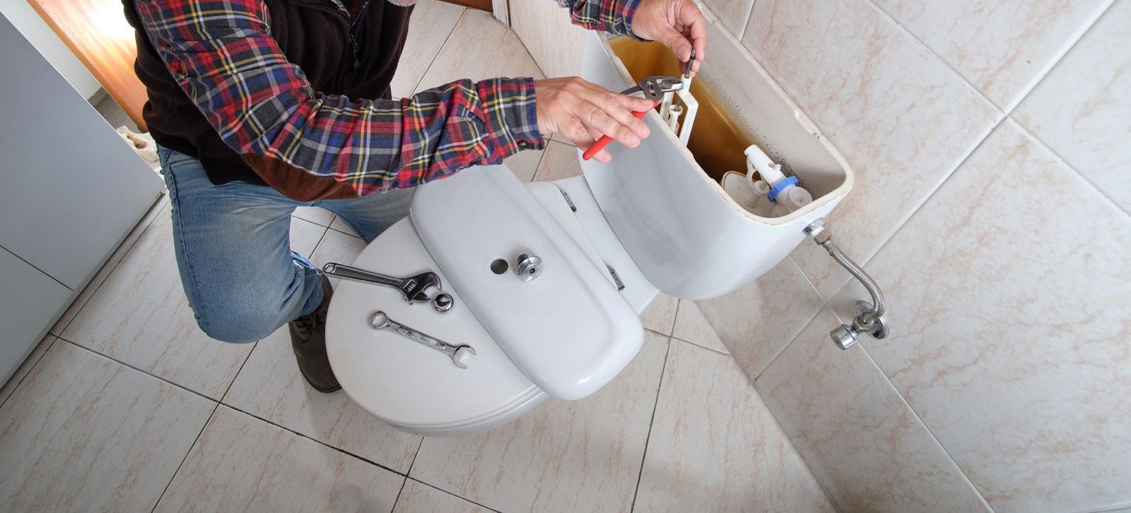 Person repairing a toilet in a bathroom, using tools to work on the tank.