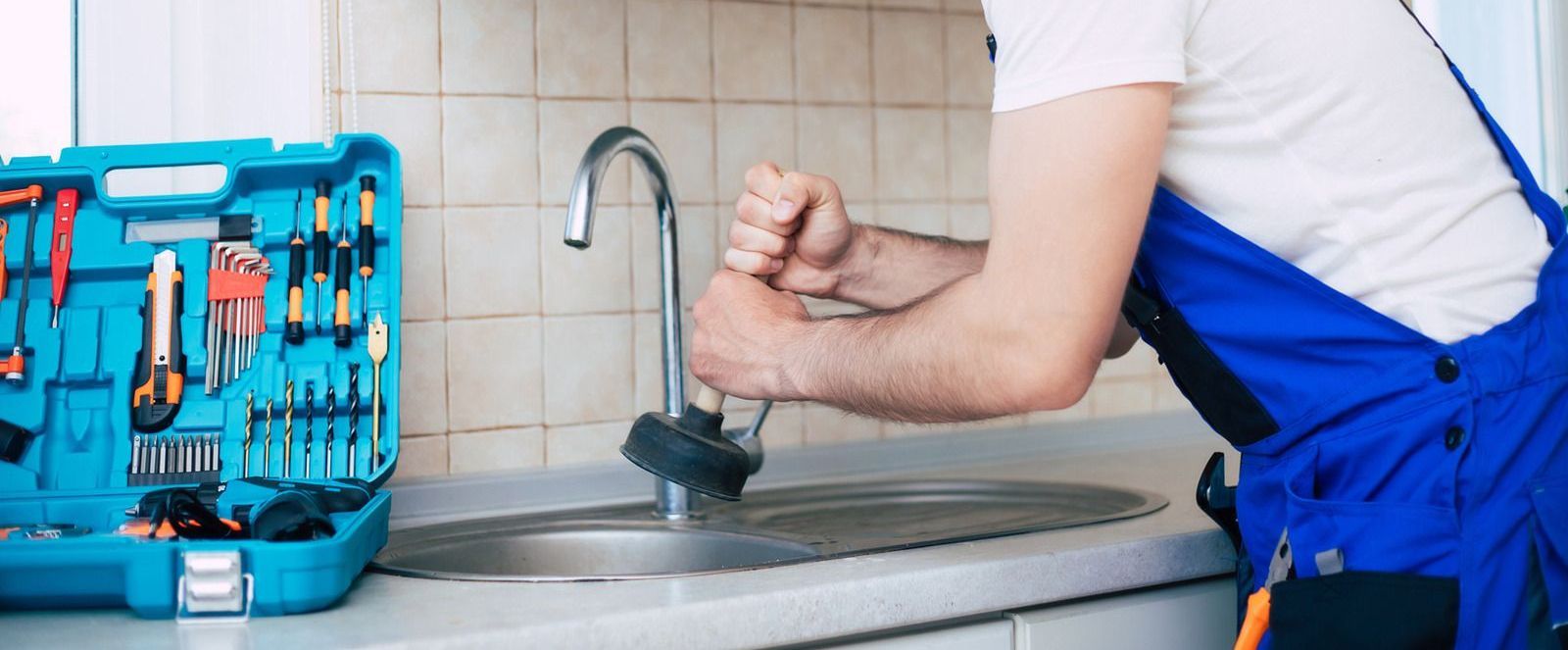 Plumber using a plunger on a kitchen sink. Tools sit in a blue toolbox. He's wearing blue overalls.