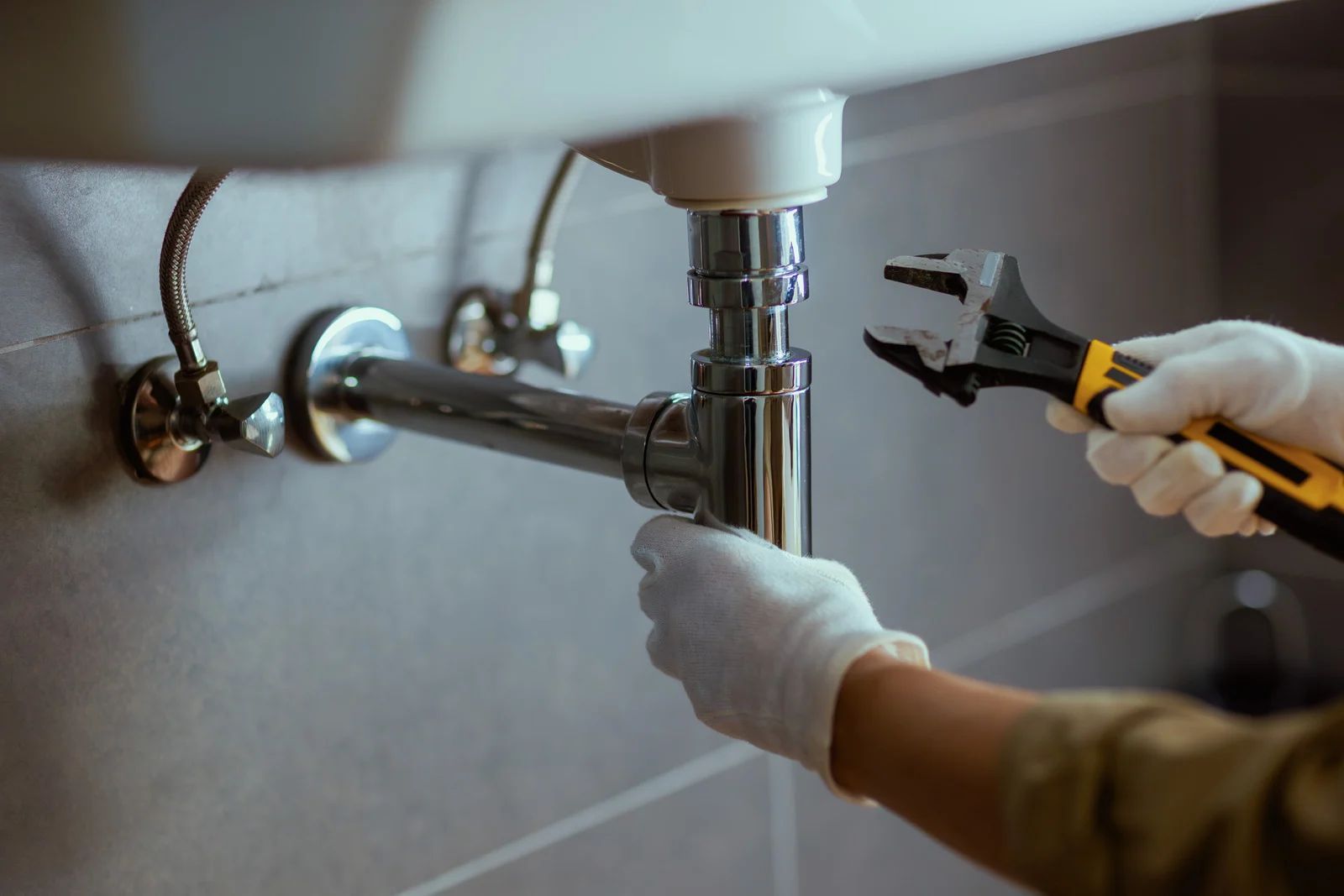 Plumber in gloves using wrench on bathroom sink pipes.