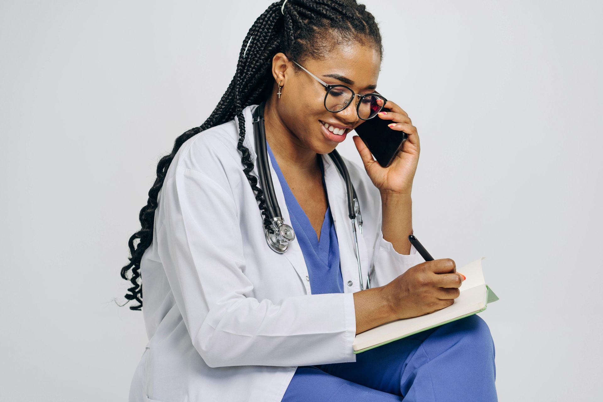 A female doctor is sitting on the floor talking on a cell phone while writing in a notebook.