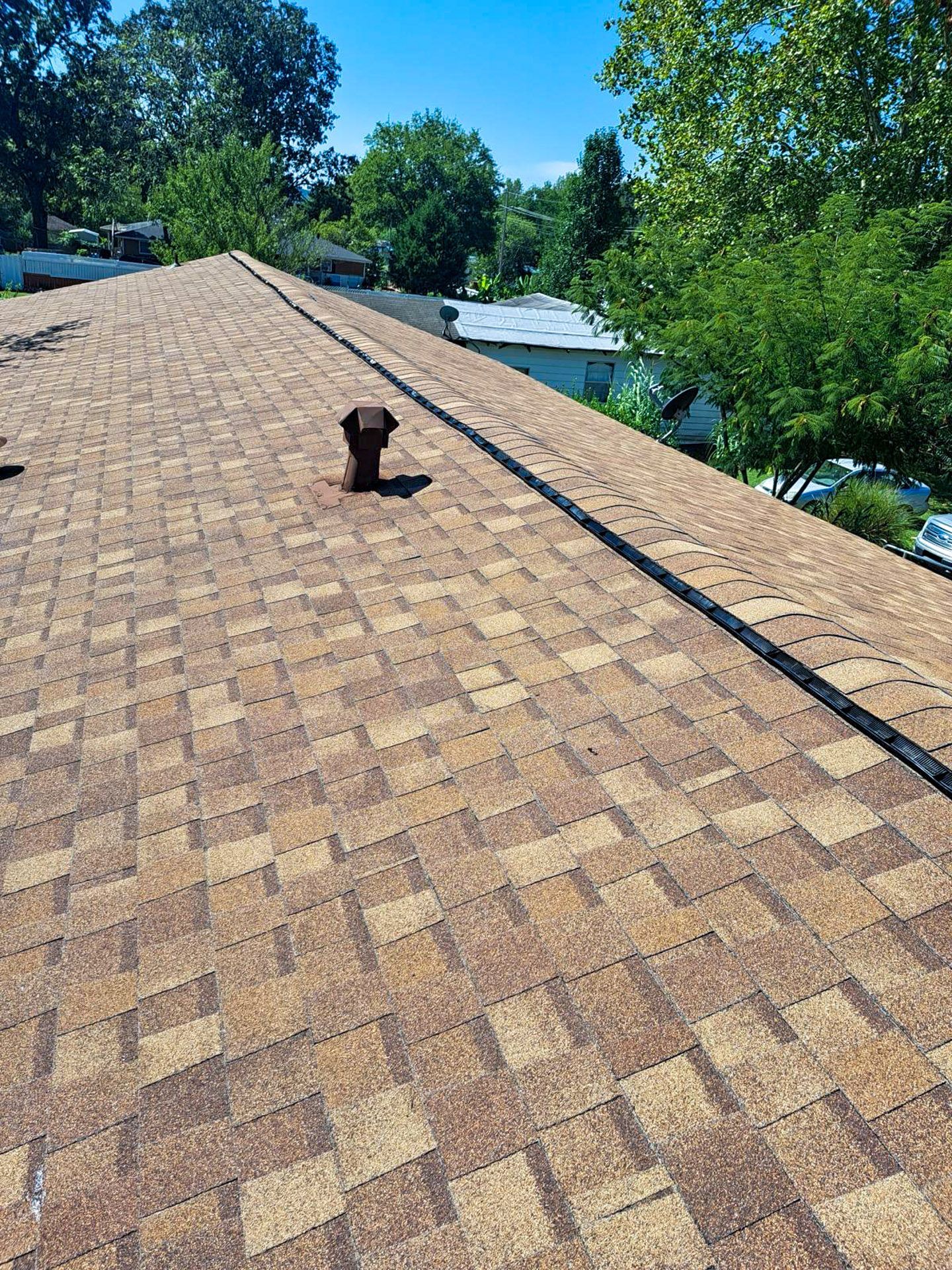 Brown asphalt shingle roof on a sunny day with a vent pipe and trees in the background.