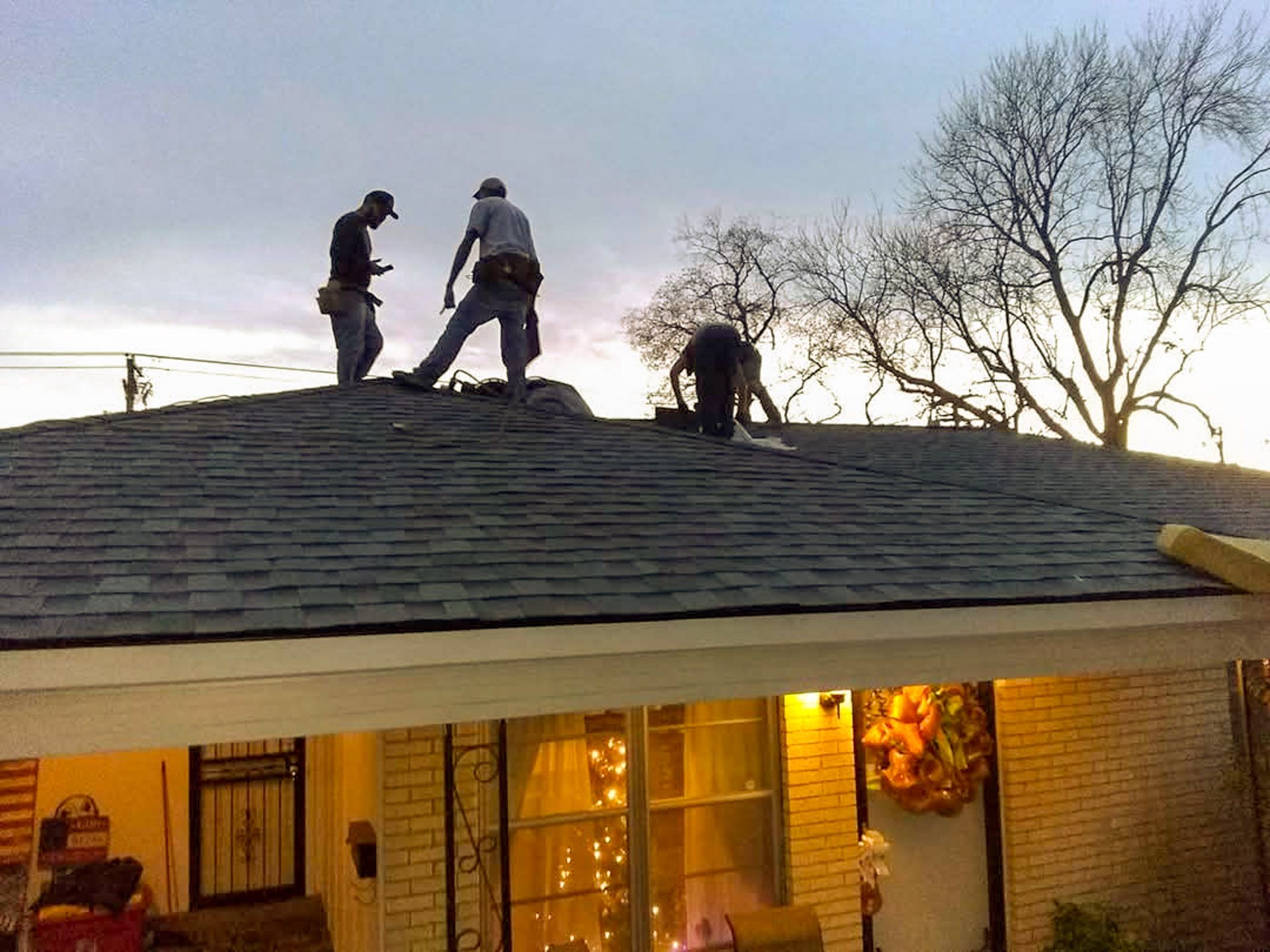 Three workers on a roof, near twilight. One looks at his phone, another stands, and one crouches near the shingles.