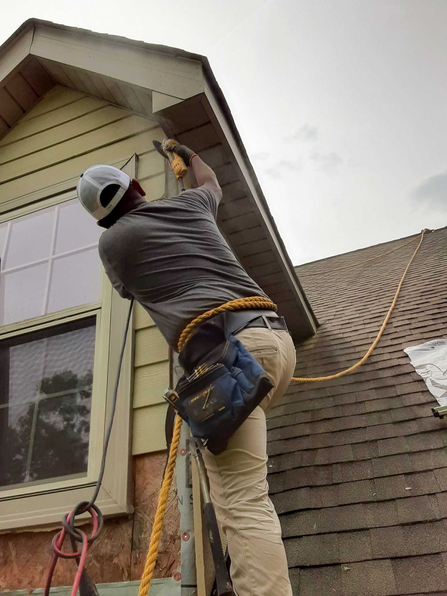 Person on ladder, working on house exterior with power tool.