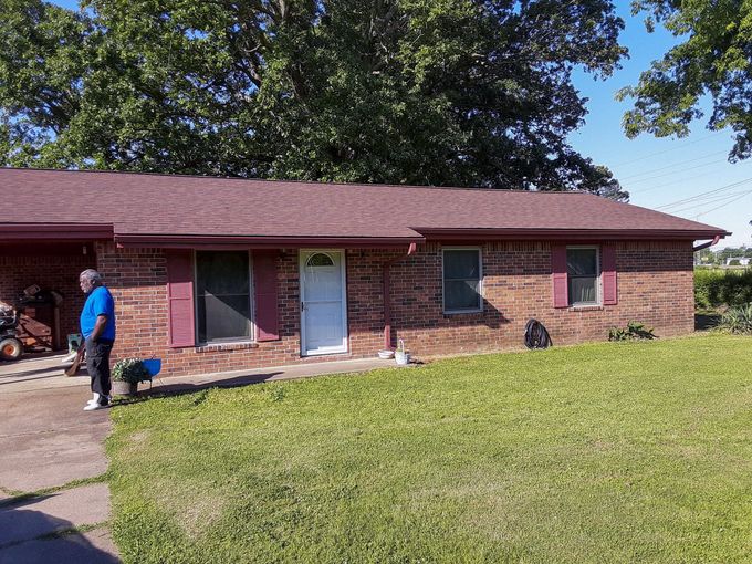 Red brick house with person and dog on the lawn. Brown roof, white door, and red shutters. Green grass and trees.