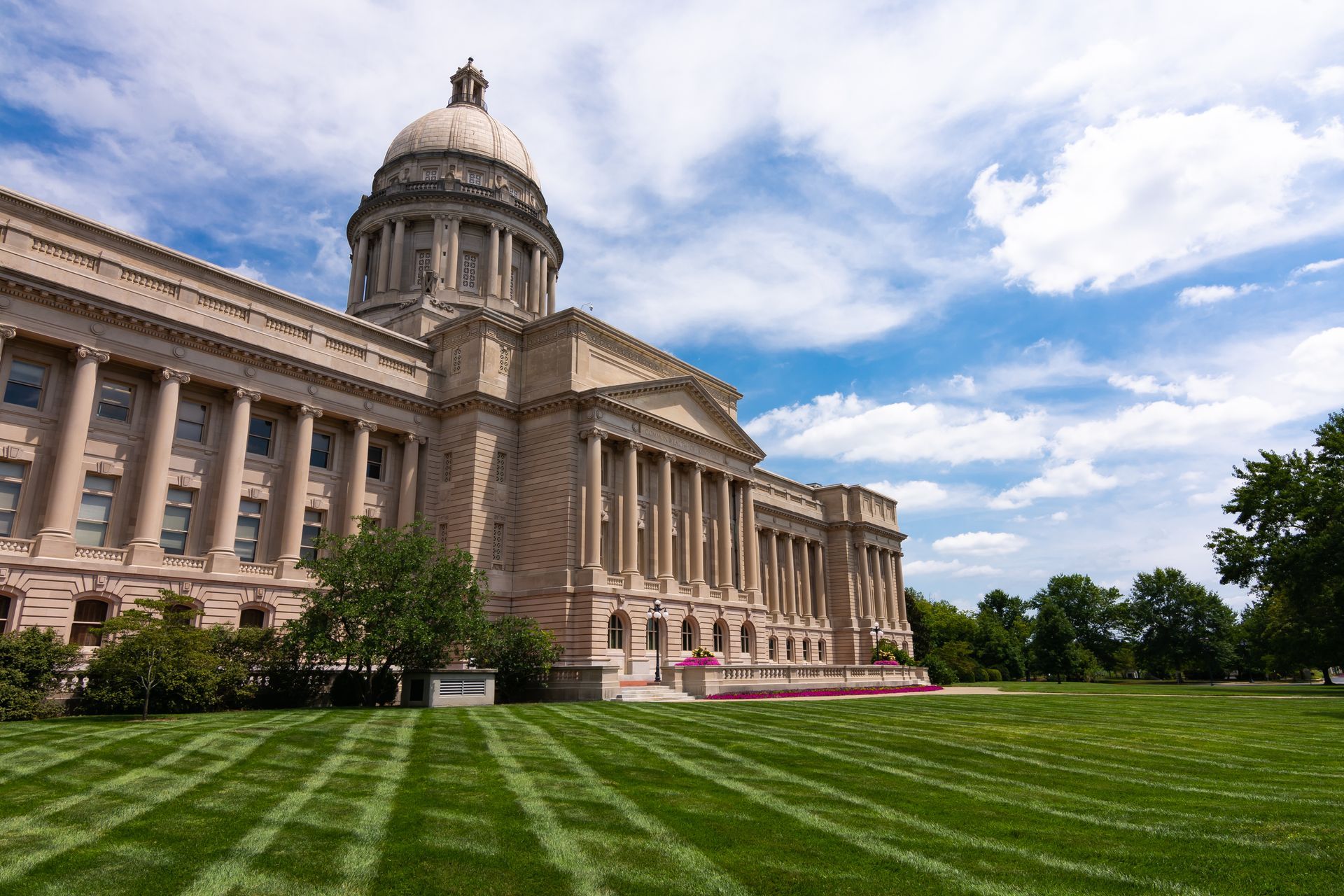 State capitol building with a domed tower, stone columns, and a green lawn under a partly cloudy sky